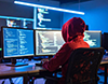 複数のモニターにプログラミングコードを表示させ、作業をする赤いパーカーの人物。